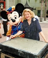 At Disney's MGM Studios, Barbara Taylor Bradford smiles after placing her hands in a cement mold as part of the 'Walk of Fame' exhibit, while Mickey Mouse playfully looks on.